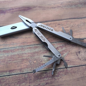 May include: A silver-toned stainless steel multi-tool featuring needle-nose pliers and various fold-out implements. The handle is engraved with the text BUILT BY DAD and the date 17.07.2026. The tool is partially open, displaying a serrated blade.