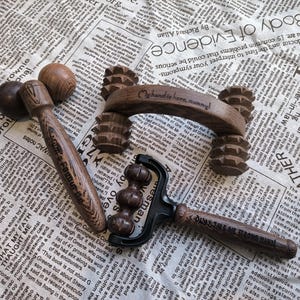 Op de afbeelding: Drie houten massagegereedschappen. De eerste is een bruine houten roller met een handvat. De tweede is een bruine houten roller met een handvat en spikes. De derde is een bruine houten roller met een handvat en een zwart metalen frame.
