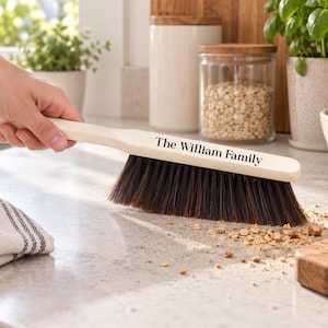 May include: A light-coloured wooden crumb brush with dark brown bristles. The handle features the black text The William Family printed on the side. The brush is shown sweeping small crumbs off a light-coloured countertop surface.