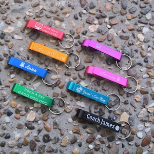May include: A collection of colourful bottle openers with personalised engravings. The openers are made of metal and have a key ring attached. Some of the engravings include names, phrases, and a phone number.