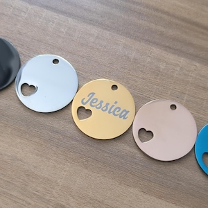 May include: Five round metal tags with a heart cut out. The tags are in black, silver, gold, rose gold, and blue. The gold tag has the name "Jessica" engraved on it.