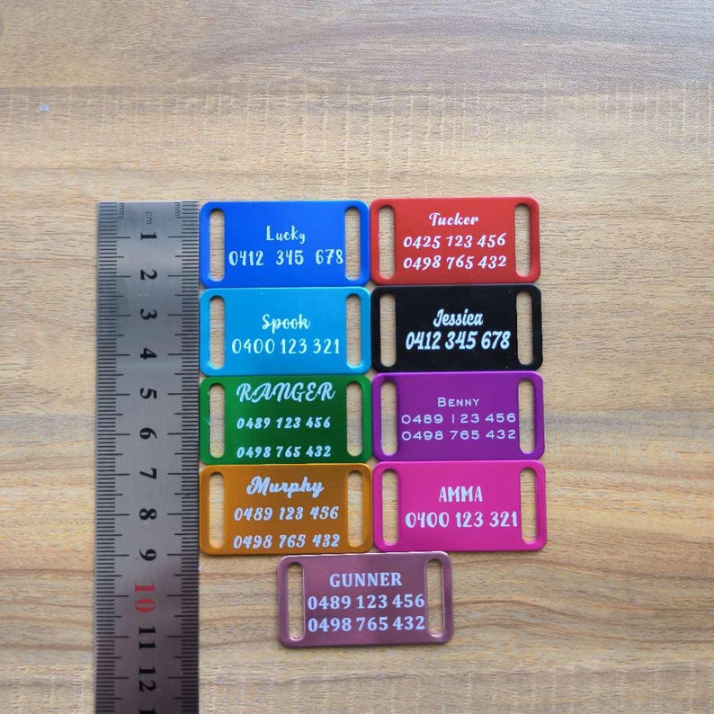 May include: Nine colorful metal dog tags with engraved names and phone numbers. The tags are arranged in a row on a wooden surface. The tags are approximately 2 inches long and 1 inch wide.