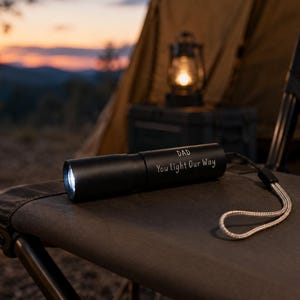 May include: A black cylindrical torch with a white wrist strap. The body of the torch features the engraved text "DAD" and "You light Our Way" in a simple font. The torch is illuminated at the front lens, emitting a bright white light.