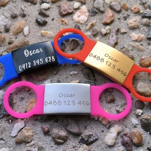 May include: Three dog ID tags with different colored silicone rings. The tags are engraved with the name "Oscar" and a phone number. The tags are blue, pink, and gold.