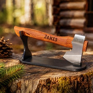 Può includere: Una piccola accetta con testa in metallo argentato lucido e manico in legno liscio con il nome JAMES inciso. L'accetta poggia orizzontalmente su un espositore in metallo nero con due supporti verticali che sostengono il manico e la lama.