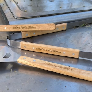 Puede incluir: Un juego de tres herramientas para asar a la parrilla con mangos de madera. Los mangos están grabados con el texto "[Name]'s Family Kitchen". Las herramientas incluyen una espátula, unas pinzas y un tenedor.