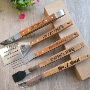 May include: A collection of personalised barbecue tools with wooden handles and stainless steel heads. The set includes tongs, a spatula, a fork, a basting brush, and a knife. Text on the tools includes "EST.2025", "Lin's Kitchen", "BBQ Masters", "Colin's BBQ", and "No. 1 Dad".