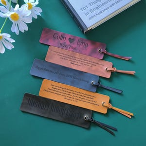 May include: Several leather bookmarks in different colours, each with engraved text and a tassel. The bookmarks are displayed on a green surface, with a book and flowers in the background. The text includes names and dates.