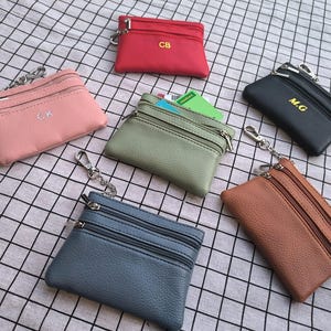 May include: A selection of small, zipped coin purses in various colours, including pink, red, green, black, blue, and brown. Each purse has a key ring and some feature initials. The purses are arranged on a checkered fabric.