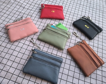 Puede incluir: Una colección de pequeños monederos con cremallera en varios colores, incluyendo rosa, rojo, verde, negro, azul y marrón. Cada monedero tiene un llavero y algunos presentan iniciales. Los monederos están dispuestos sobre una tela a cuadros.