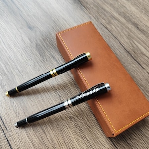 May include: Two black pens with gold and silver detailing, one engraved with the name "Amanda", are placed on a wooden surface beside a brown leather pen case. The pens have a sleek, professional design.