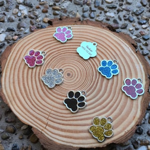 May include: Assortment of seven silver paw print dog tags with glitter accents. The tags are in various colors including pink, blue, silver, black, and gold. One tag has the name "Tucker" and a phone number engraved on it.