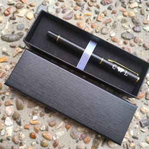 Puede incluir: Un bolígrafo negro con detalles dorados y las iniciales "C & L" grabadas, en una caja de regalo negra.