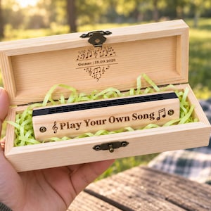 Könnte beinhalten: Ein helles Mundharmonika-Etui aus Holz mit Metallverschluss, das eine Mundharmonika aus Holz enthält, graviert mit dem Text "Play Your Own Song" und einem Notensymbol. Der Innendeckel zeigt Noten und den Text "Quinne | 18.09.2026".