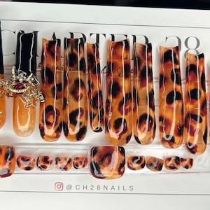 May include: Set of 10 false nails with a leopard print design. The nails are in various shapes and sizes, with some being longer and more oval-shaped, while others are shorter and more square-shaped. The nails are a light brown color with black spots.