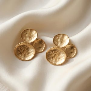 May include: A pair of gold-toned earrings, each featuring three circular, hammered-texture discs of varying sizes arranged in a cluster. The metal surface has a matte, reflective finish with irregular indentations across the rounded shapes.