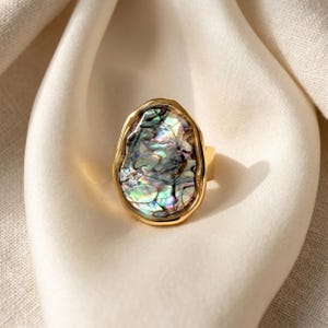 May include: A gold-toned ring featuring an iridescent, oval-shaped stone. The stone displays a mix of colors, including green, blue, and purple, set within a textured gold frame. The ring's band is a simple, polished gold.