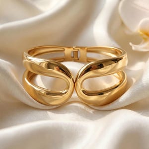 May include: A polished gold-tone hinged cuff bracelet featuring a wide, open-work design with two symmetrical, rounded, fluid-shaped loops that meet at the center. The metal surface is smooth and reflective, showcasing a continuous, curved silhouette.
