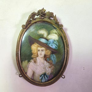 May include: Oval portrait painting in a gold-toned frame. The painting depicts a woman with blonde curly hair wearing a hat with feathers and a blue ribbon. The background is green. The frame has decorative floral elements at the top and small hanging loops.