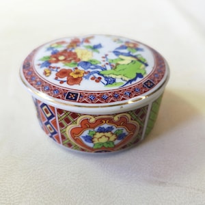 May include: A small, round ceramic trinket box with a lid. The lid and sides are decorated with colourful floral and geometric patterns in red, blue, yellow, and green. The box has a gold trim and sits on a white surface.