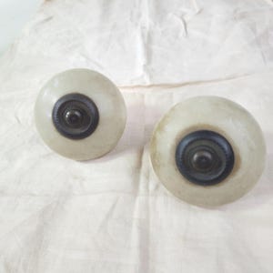 Puede incluir: Dos pomos de armario redondos de color blanquecino con un centro circular oscuro. Los pomos tienen un aspecto ligeramente envejecido, lo que sugiere un estilo vintage o antiguo. Probablemente se utilizan para cajones o puertas de armarios.
