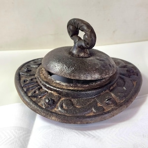 Peut inclure: Un couvercle circulaire en fonte sombre et patinée, doté d'une poignée centrale en forme de boucle et d'une bordure décorative en relief avec des motifs de volutes répétitifs. La surface métallique présente une texture vieillie.