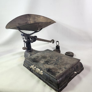 May include: An antique Acme scale with a large, curved metal tray. The scale is dark grey with visible wear and tear, and the word "Acme" is printed on the base. Several weights are placed next to the scale.