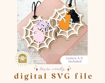 Può includere: Due ornamenti in legno a forma di ragnatela tagliati al laser con un ragno e una lettera su ciascuno. Le ragnatele sono bianche con linee nere, i ragni sono neri e viola, e le lettere sono arancioni e rosa. Il testo "Lettere A-Z incluse" è in basso nell'immagine.