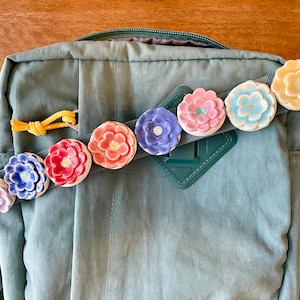 Puede incluir: Una mochila verde con una fila de abalorios de flores de cerámica de colores unidos a la correa. Los abalorios son en tonos de rosa, rojo, azul, amarillo y morado.