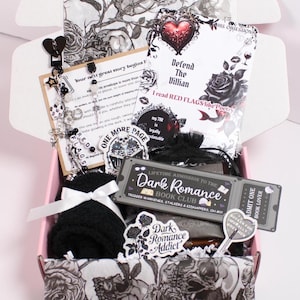 May include: A pink box filled with dark romance-themed items. Includes a book titled "Defend The Villian", stickers, a bookmark, a black knitted item, and a ticket that reads "Dark Romance Book Club". The box is decorated with black and white floral patterns.