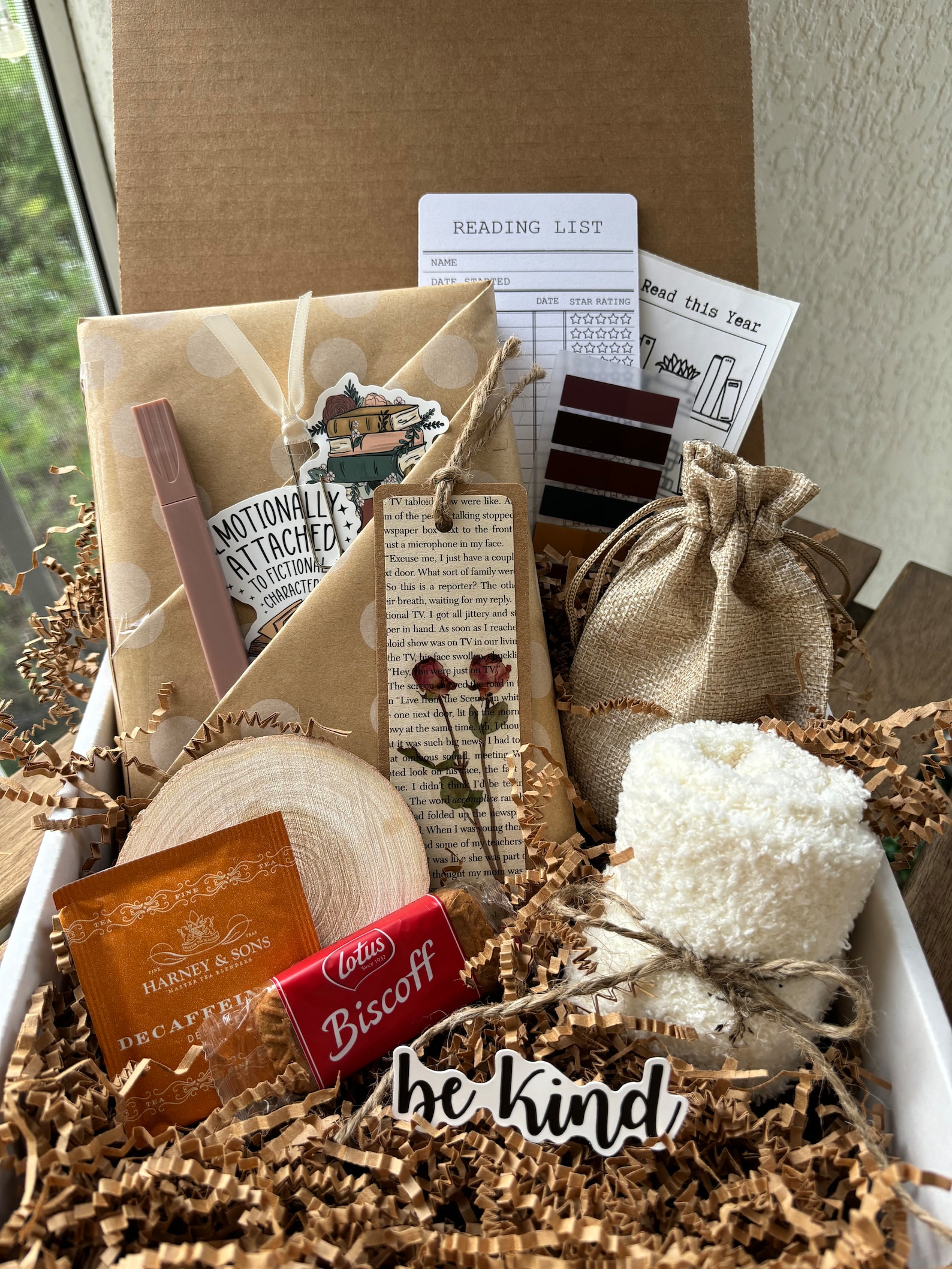 A cozy gift box filled with a wrapped book, a "be kind" sticker, tea, Biscoff cookies, fluffy socks, a natural wood slice, bookmarks, and a reading list.