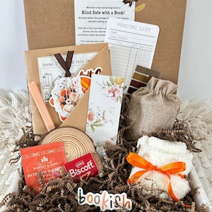 May include: A brown box filled with a bookish gift set. The box contains a book, a brown envelope, a wooden coaster, a bag of tea, a package of cookies, a pair of socks, a small burlap bag, and a reading list. The box is decorated with fall-themed stickers and paper.