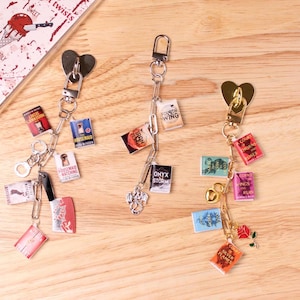 May include: Three keychains with miniature book charms, a cleaver, and heart-shaped accents. The keychains are silver-toned and feature small book covers with titles like "Fourth Wing" and "Housemaid Watching". The background includes text that reads "Sweet twists but deadly".