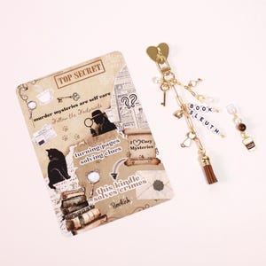 May include: A beige scrapbook-style card with text and illustrations, including a cat, books, and the words "TOP SECRET." A gold keychain with charms, including a tassel, key, and the word "BOOK SLEUTH." A coffee cup charm is also present.