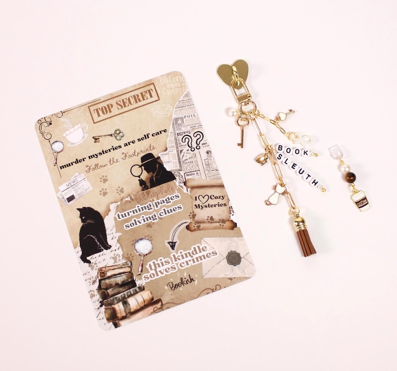 May include: A beige scrapbook-style card with text and illustrations, including a cat, books, and the words "TOP SECRET." A gold keychain with charms, including a tassel, key, and the word "BOOK SLEUTH." A coffee cup charm is also present.