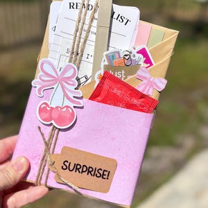 Blind Date with a Book mystery gift box for readers, offering a handpicked book from genres like romantasy, dark romance, or sci-fi. The book is beautifully gift-wrapped with clues, along with extra goodies such as herbal tea, bookish stickers, annotation tabs, a highlighter, book tracker, and a library-style reading list. Ideal for booktok enthusiasts, book lovers, and those who enjoy a thrilling or romantic surprise reading experience.