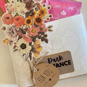 May include: A 'Blind Date with a Book' gift set, featuring a book, tea, and other small gifts. The set is decorated with floral stickers, tags, and twine. The text on the tag says 'For You' and 'Dark Romance'.