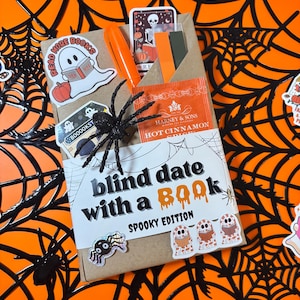 May include: A brown paper-wrapped book labeled Blind Date with a Book Spooky Edition, decorated with ghost and spider stickers, a black plastic spider, an orange highlighter, and a packet of Harney & Sons Hot Cinnamon Spice tea.