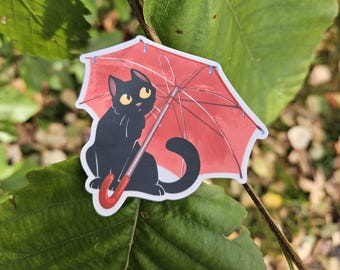 May include: A sticker of a black cat sheltering under a red umbrella. The cat has yellow eyes and is holding the umbrella. The umbrella has a red handle and a blue trim. The sticker is on a green leaf.