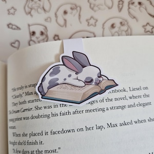 May include: A bookmark featuring a cartoon rabbit with grey spots and ears, resting on a book. The rabbit is depicted sleeping on the book's pages. The bookmark is white with a small tab to insert into a book.