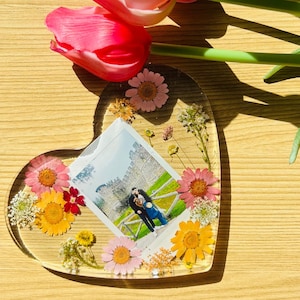 Pode incluir: Um descanso de copo de resina em forma de coração com flores prensadas e uma foto de um casal em frente a um castelo. O descanso de copo é transparente e as flores são de várias cores, incluindo rosa, amarelo e branco.
