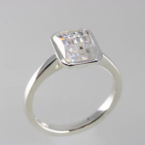Peut inclure: Une bague argentée dotée d'une pierre transparente rectangulaire à facettes, montée dans un serti clos lisse. L'anneau est poli et plein, avec une surface propre et réfléchissante qui met en valeur la clarté de la pierre et la finition du métal.