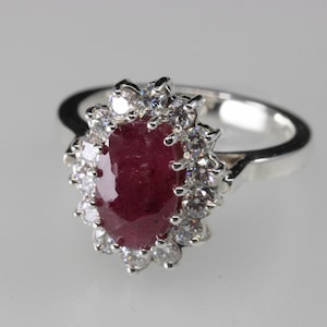 Puede incluir: Anillo de plata con una piedra roja oscura ovalada en el centro, rodeada por un halo de piedras claras más pequeñas. La banda es de plata pulida. Una joya elegante, ideal para ocasiones especiales.