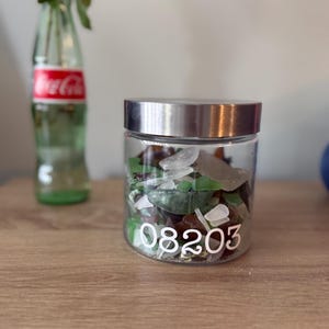 May include: A clear glass jar with a silver-toned metal lid, filled with assorted pieces of smooth, multi-colored sea glass. The front of the jar features the white printed numbers 08203. The container is cylindrical with a flat base.