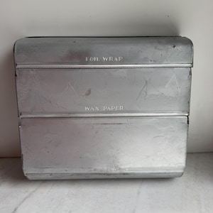 May include: A vintage, rectangular metal kitchen organizer with the words "FOIL WRAP", "WAX PAPER", and "PAPER TOWELS" embossed on the front. The organizer is silver and has a slightly worn appearance, suggesting age and use.