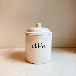 Puede incluir: Un tarro de cerámica blanco con tapa y un pequeño pomo. La palabra "edibles" está escrita en negro en la parte delantera. El tarro es cilíndrico y tiene una línea negra en la parte superior.