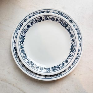 Puede incluir: Dos platos de cerámica blanca apilados uno encima del otro. Los platos tienen un patrón floral azul alrededor del borde. Los platos están sobre una superficie de mármol blanco y gris.