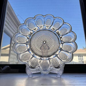 May include: A clear glass egg plate with a raised, textured center and 12 individual egg-shaped depressions around the edge. The plate is on a white stand.