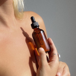 以下が含まれることがあります： 黒いドロッパーが付いた茶色のガラス瓶で、透明な液体が入っています。瓶は人の手で持たれています。