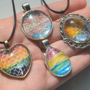 Pode incluir: Quatro colares com diferentes formas de pingentes e designs de resina em cores do arco-íris. Os pingentes são em forma de coração, círculo, oval e lágrima. Cada pingente tem uma moldura de metal prateado.